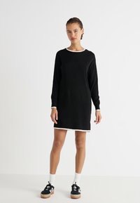 Zwarte gebreide jurk met lange mouwen en een ronde halslijn, voorzien van witte afwerking bij de manchetten en dezoom, gecombineerd met zwarte sneakers en witte sokken.