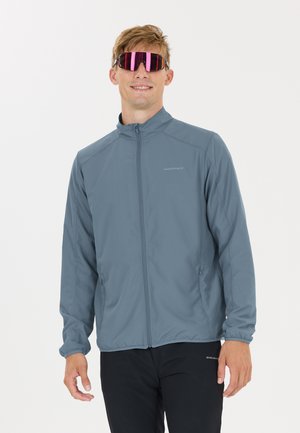 Mann trägt graue Jacke mit Reißverschluss und schwarze Hose, dazu reflektierende rote Sportsonnenbrille, lächelt vor weißem Hintergrund.