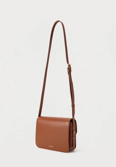 Καφέ δερμάτινη τσάντα ώμου με ρυθμιζόμενο λουρί και κλείσιμο με καπάκι, μινιμαλιστικός σχεδιασμός και μικρό χρυσό λογότυπο στην πρόσοψη.