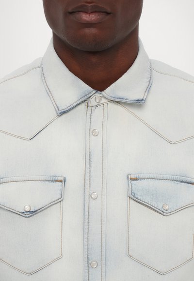 Lyseblå denimskjorte med spidse kraver, trykknapper, to brystlommer med klapper og tan-stikninger, der fremhæver sømmene.