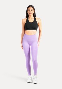 Schwarzer Sport-BH mit rundem Ausschnitt, kombiniert mit hoch taillierten, figurbetonten Lavendel-Leggings. Das Model trägt weiße Sportschuhe.