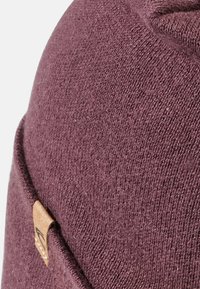 Maroon beanie made of soft knit material. Features a textured surface and a small brown leather tag on the side. Round shape.