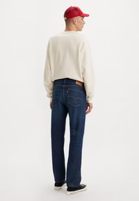 Suéter blanco de mangas largas, vaqueros rectos de color azul oscuro con costuras naranjas y zapatillas negras. Gorra roja con parte trasera de malla.