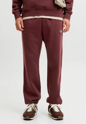 Personne portant un pantalon de survêtement bordeaux, un sweat assorti et des baskets marron et blanches, debout devant un fond uni.