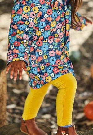 Enfant portant une veste fleurie colorée, un pantalon jaune vif et des bottes brunes avec un motif de renard, en équilibre sur une souche en bois en plein air.