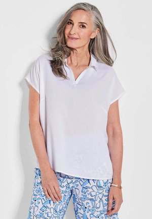 Blouse blanche à manches courtes avec un col en V et en tissu doux ; portée avec un pantalon bleu à motifs présentant des motifs floraux bleus.