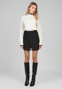 Un pull en tricot crème, une jupe noire rayée avec un ourlet en dentelle, et des bottes noires jusqu'aux genoux complètent la tenue. Des détails de texture et de design subtils sont présents.