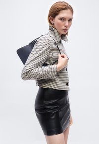 Γυναίκα που φοράει τζάκετ με καρό σχέδιο και μαύρη δερμάτινη φούστα, κρατώντας μια μαύρη τσάντα στον ώμο, στέκεται μπροστά σε ένα απλό φόντο.