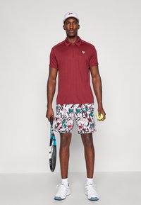 Bordo polo majica s kratkim rukavima, uzorkane kratke hlače u bijeloj, ružičastoj i zelenoj boji, drži tenisku lopticu i žuti teniski reket.