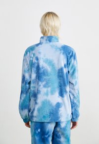 Fleecejakke i lyseblå med et tie-dye mønster i ulike nyanser av blått. Den har høy krage og lange ermer.