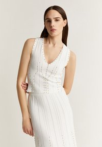 Hvid strikket to-delt outfit med V-hals, ærmeløs top, lodret ribbet tekstur og guldbelægning langs sømmene.