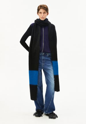 Person, die einen langen schwarz-blauen Schal, ein navyfarbenes Zip-Hoodie, weite blaue Jeans und schwarze Fransen Schuhe trägt, steht mit einer Hand in der Hüfte.