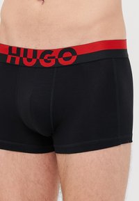 Schwarze Boxershorts mit einem roten Gummibund mit "HUGO"-Logo. Glattes Material, taillierte Passform und kurze Beinlänge.
