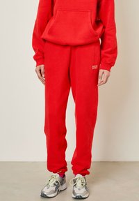 Sarkana flīsa kapuce un atbilstoši sporta bikses, abām ir brīvs piegriezums un priekšējais kangaroo kabata; logo detalizācija biksēs.