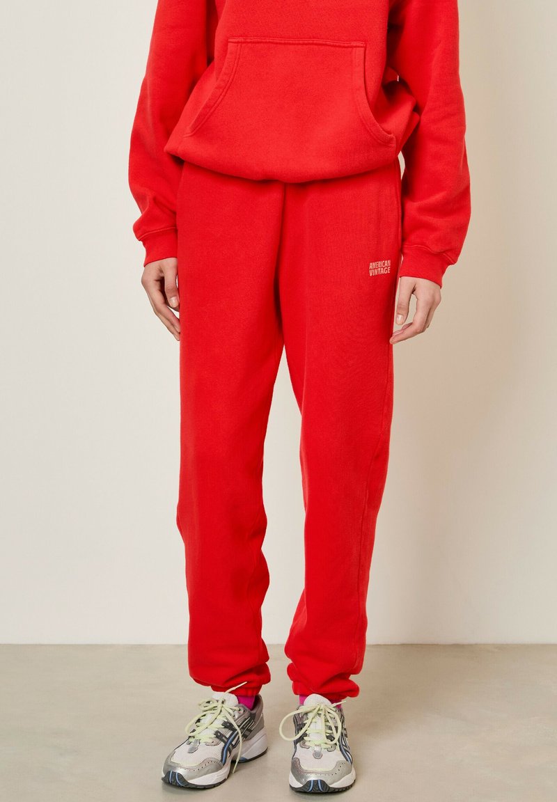 Sarkana flīsa kapuce un atbilstoši sporta bikses, abām ir brīvs piegriezums un priekšējais kangaroo kabata; logo detalizācija biksēs.