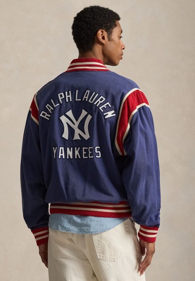 Άνδρας που φοράει μπλε και κόκκινο μπουφάν Ralph Lauren Yankees, ανοιχτό μπλε πουκάμισο και μπεζ παντελόνι, εικονιζόμενος από πίσω σε ουδέτερο φόντο.