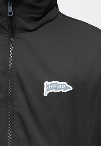 Veste noire avec une fermeture éclair frontale et un petit écusson en forme de drapeau portant le logo "HUGO" sur la poitrine.