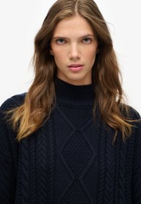 Suéter de punto azul marino con cuello alto, con patrones texturizados de cable y detalles en forma de diamante. Material suave y grueso con una apariencia acogedora.