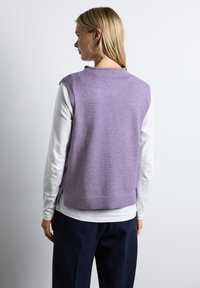 Ljuslila ärmlös tröja med ribbad nederkant, bärs över en vit långärmad skjorta med en enkel rund halsringning.