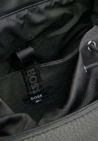 Interior de bolso de cuero negro con forro de tela. Cuenta con una etiqueta tejida con la palabra "BOSS" y bolsillos minimalistas. Textura suave y diseño elegante.