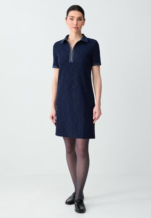 Kobieta ubrana w granatową, krótki rękaw, teksturowaną sukienkę z zamkiem przy kołnierzu, czarne przezroczyste rajstopy i czarne półbuty, stojąca na białym tle.