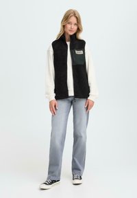 Vrouw in een zwart fleece vest over een witte sweatshirt, lichte grijze spijkerbroek en zwarte lage sneakers, staande tegen een effen achtergrond.