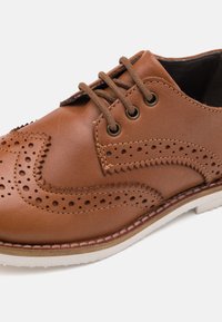 Scarpa oxford in pelle marrone con dettagli perforati, punta tonda, sei occhielli e suola in gomma bianca a contrasto con cuciture a vista.
