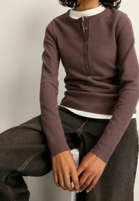 Osoba ubrana w zapinaną na guziki, śliwkową, długorękawową koszulę na białej bluzce, w ciemnych dżinsach, siedząca z rękami spoczywającymi na kolanach.