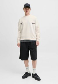 Creme sweatshirt med logo, sorte shorts, sorte snørebåndssko og hvide sokker med tekst. Afslappet outfit, ensfarvede farver, løs pasform.