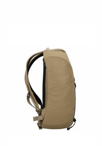 Mochila compacta marrón con correas de hombro acolchadas, bolsillo lateral y correas ajustables, mostrada en vista lateral sobre un fondo blanco.