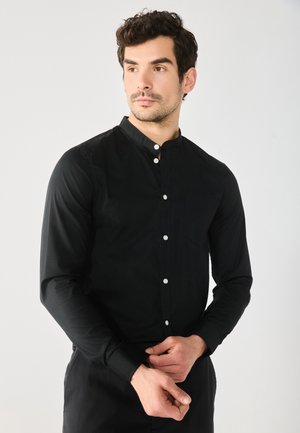 Camicia nera con colletto alto, maniche lunghe, bottoni bianchi e tasca frontale. Tessuto liscio con un design aderente.