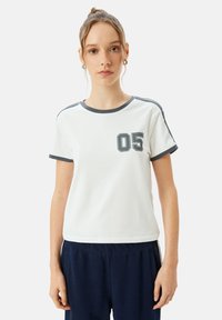 Hvid kortærmet T-shirt med mørkegrå kant og tallet "05" i en firkant på venstre bryst. Fremstillet af bomuldsstof, afslappet pasform.