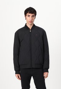 Black quilted bomber jacket with a stand collar, featuring a zip closure, ribbed cuffs, and a small logo detail on the chest.