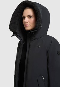 Parka negra con una gran capucha forrada en tejido texturizado. Presenta un cierre con cremallera y un logo en el pecho, con botones a presión en las mangas.