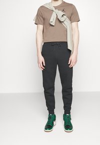 T-shirt in cotone marrone con logo nero, abbinato a pantaloni jogger neri e sneakers alte verdi con accenti neri e bianchi.