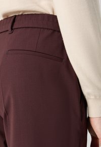 Burgundy trousers with a smooth texture, featuring a back pocket and a tailored fit. Worn with a light-colored, textured sweater.