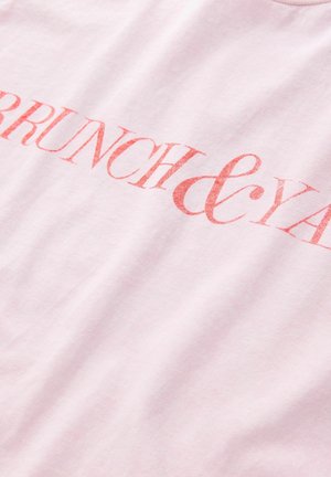 Gaiši rozā kokvilnas T-krekls ar drosmīgu, teksturētu sarkanu uzrakstu "BRUNCH & YAY." Vienkārša apaļa kakla izgriezums ar īsām piedurknēm.