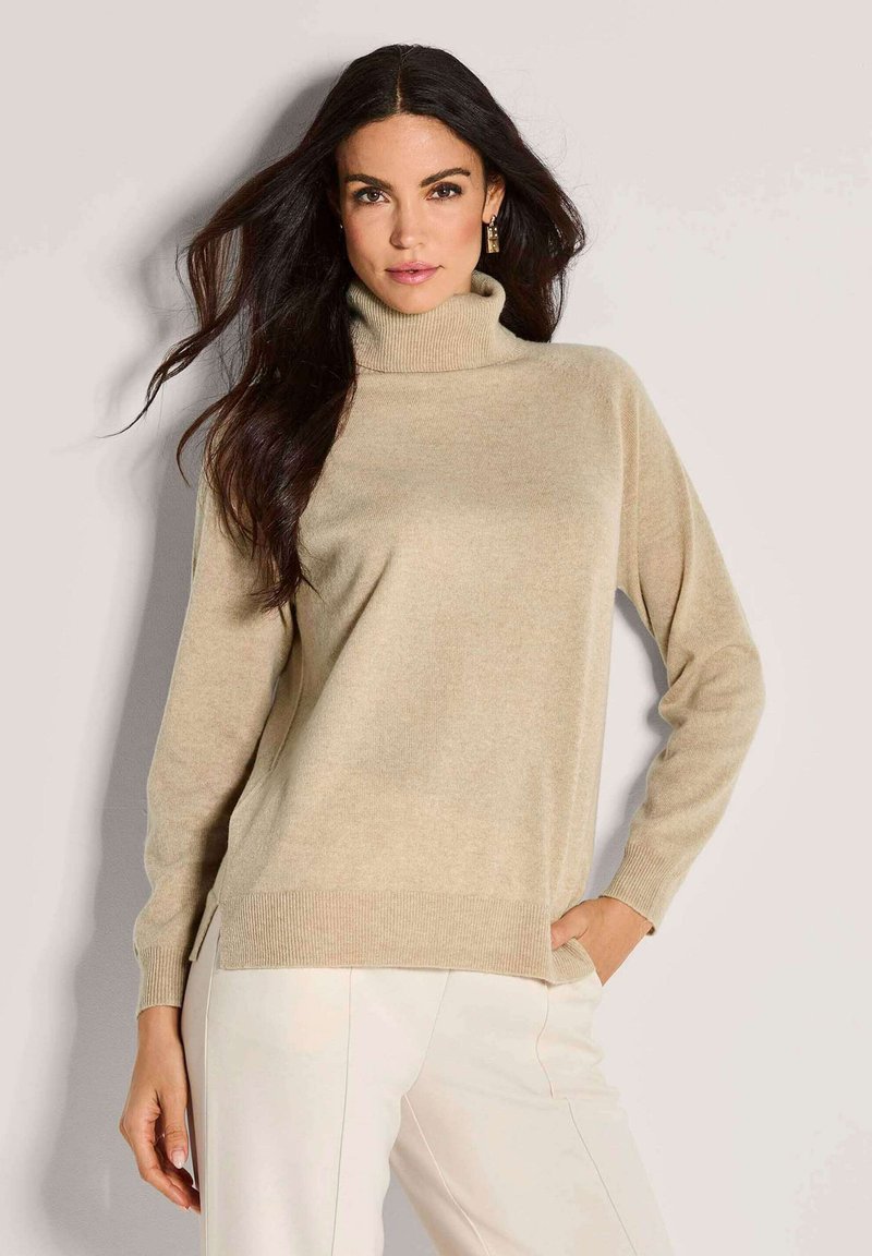 Frau mit langen dunklen Haaren, die einen beigen Rollkragenpullover und helle Cremefarbene Hose trägt, posiert vor einem schlichten hellen Hintergrund.