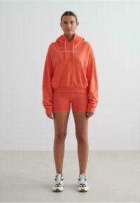 Koraalkleurige hoodie met een buidelzak en verstelbaar trekkoord, gecombineerd met bijpassende koraalkleurige shorts, allemaal gemaakt van zacht materiaal.