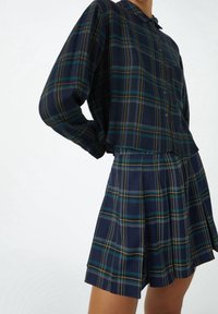 Outfit a quadri, composto da una camicia a maniche lunghe con bottoni e una gonna plissettata. Base blu scuro con strisce verdi e gialle. Tessuto morbido.