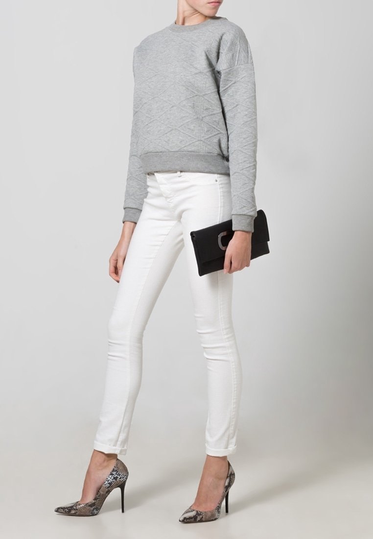 Personne portant un pull gris à motifs, un pantalon blanc ajusté, des escarpins imprimé serpent, tenant une pochette noire devant un fond uni.