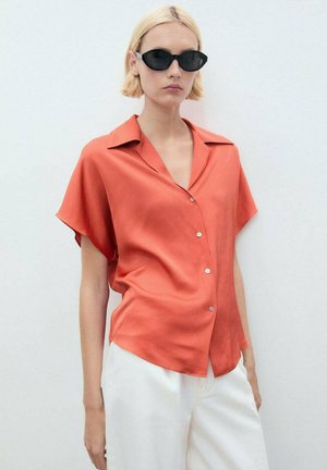 Blusa a maniche corte con bottoni in un vivace corallo, realizzata in un tessuto morbido. Presenta un colletto classico e una leggera lucentezza. Abbinata a pantaloni bianchi.