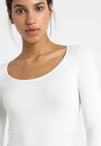 Haut à manches longues blanc avec un col arrondi, fabriqué en tissu lisse et doux, épousant étroitement le corps, avec des coutures minimales.