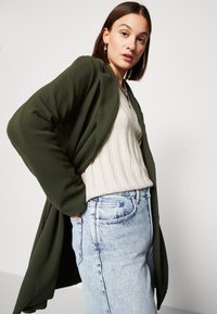 Olive green textured jacket, open front, worn over a cream ribbed sweater and light blue jeans. Details include a layered look and slouched silhouette.