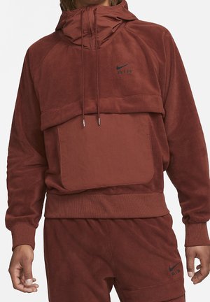 Mężczyzna ubrany w rdzawy, polarowy kaptur Nike Air oraz pasujące spodnie z przednią kieszenią i sznurkiem przy kapturze, przycięte przy szyi i talii.