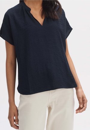 Frau trägt kurzärmeliges, strukturiertes dunkelblaues Oberteil mit V-Ausschnitt und cremefarbene, hoch taillierte Hose vor einfarbigem hellem Hintergrund.