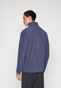 Granatowa polarowa kurtka z wysokim kołnierzem i długimi rękawami, o gładkiej fakturze i luźnym kroju, widziana od tyłu.