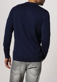 Granatowy sweter typu pullover z okrągłym dekoltem, długimi rękawami oraz ściągaczami przy mankietach i dolnym brzegu, w zestawieniu z jasnoszarymi dżinsami o subtelnym wzorze.