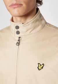 Beige jakke med høy krage, to mørke knapper, glidelås og en gul brodert logo på brystet. Teksturert stoff.
