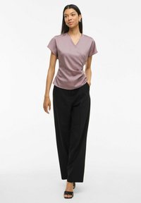 Blusa avvolgente a maniche corte di colore malva in tessuto morbido, abbinata a pantaloni neri a vita alta. Il modello è in piedi con sicurezza in tacchi neri aperti.